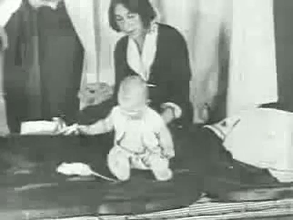 A grainy frame of silent film: a baby in a white gown sits on a dark mattress while a white rat is placed before him, and a young woman in a dark dress with a white collar watches from behind