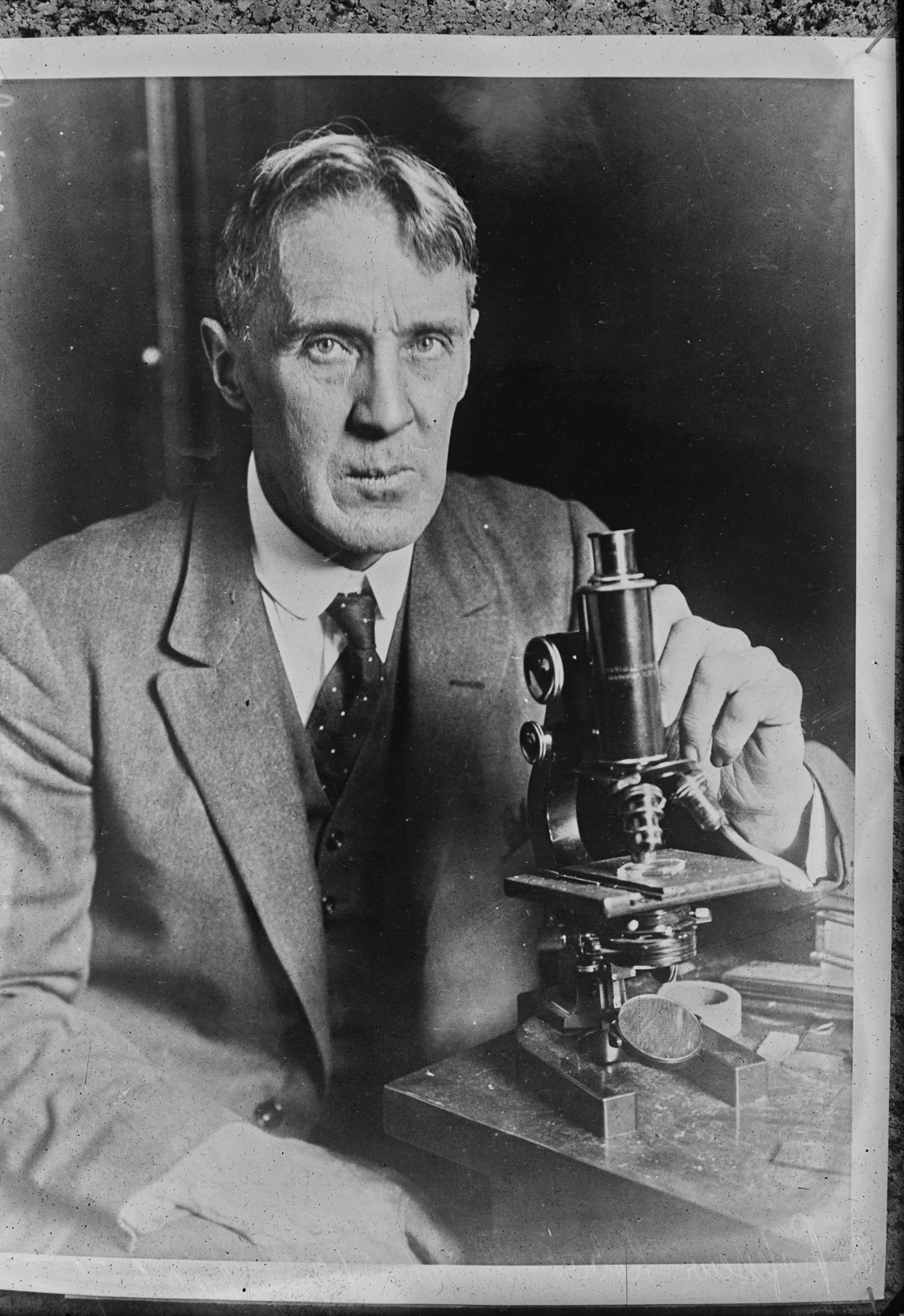 A photographic portrait of a lean grey-haired man in a tweed jacket, seated, with one hand on the focusing knob of a small brass microscope on the table beside him