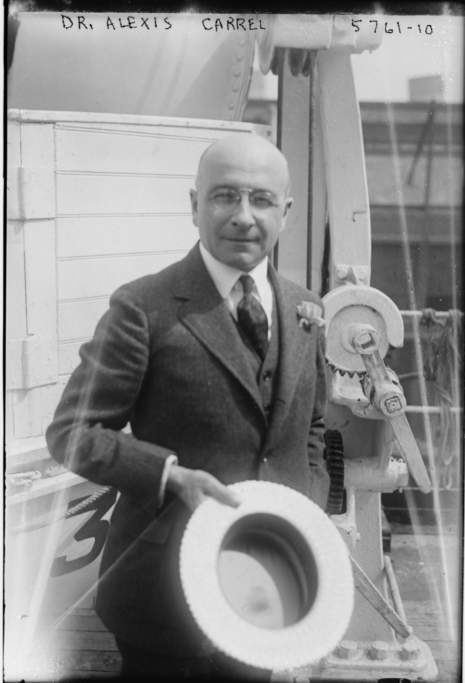A bald man in a dark three-piece suit and round spectacles stands on a ship's deck holding a straw boater, with a winch behind him
