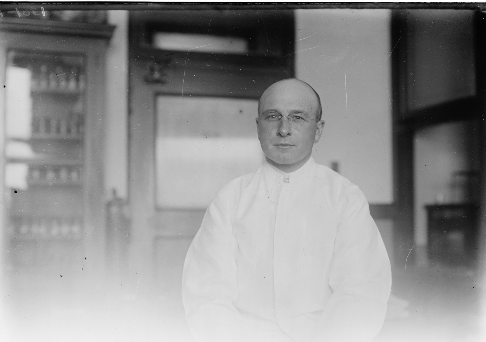 A bald man with round spectacles in a white laboratory coat, seated in front of a glass-fronted cabinet of bottles