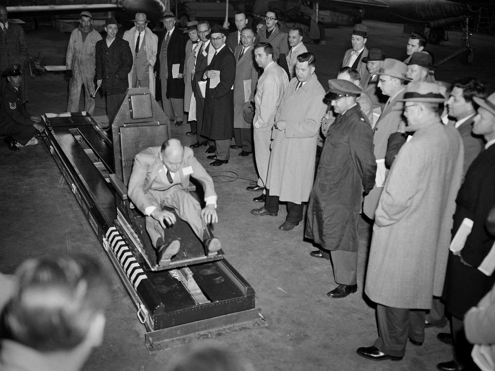 In a hangar, a man in a suit sits strapped in a seat on a low rail-mounted sled, his hands gripping the sides, while two dozen men in overcoats and hats stand round watching