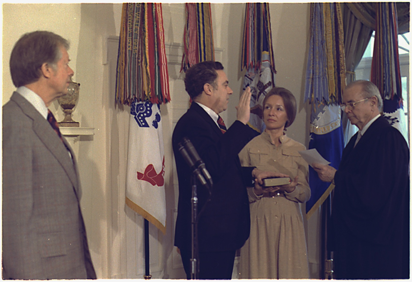 Joseph Califano raising his right hand to take the oath of office as his wife holds a Bible and President Carter watches from the left