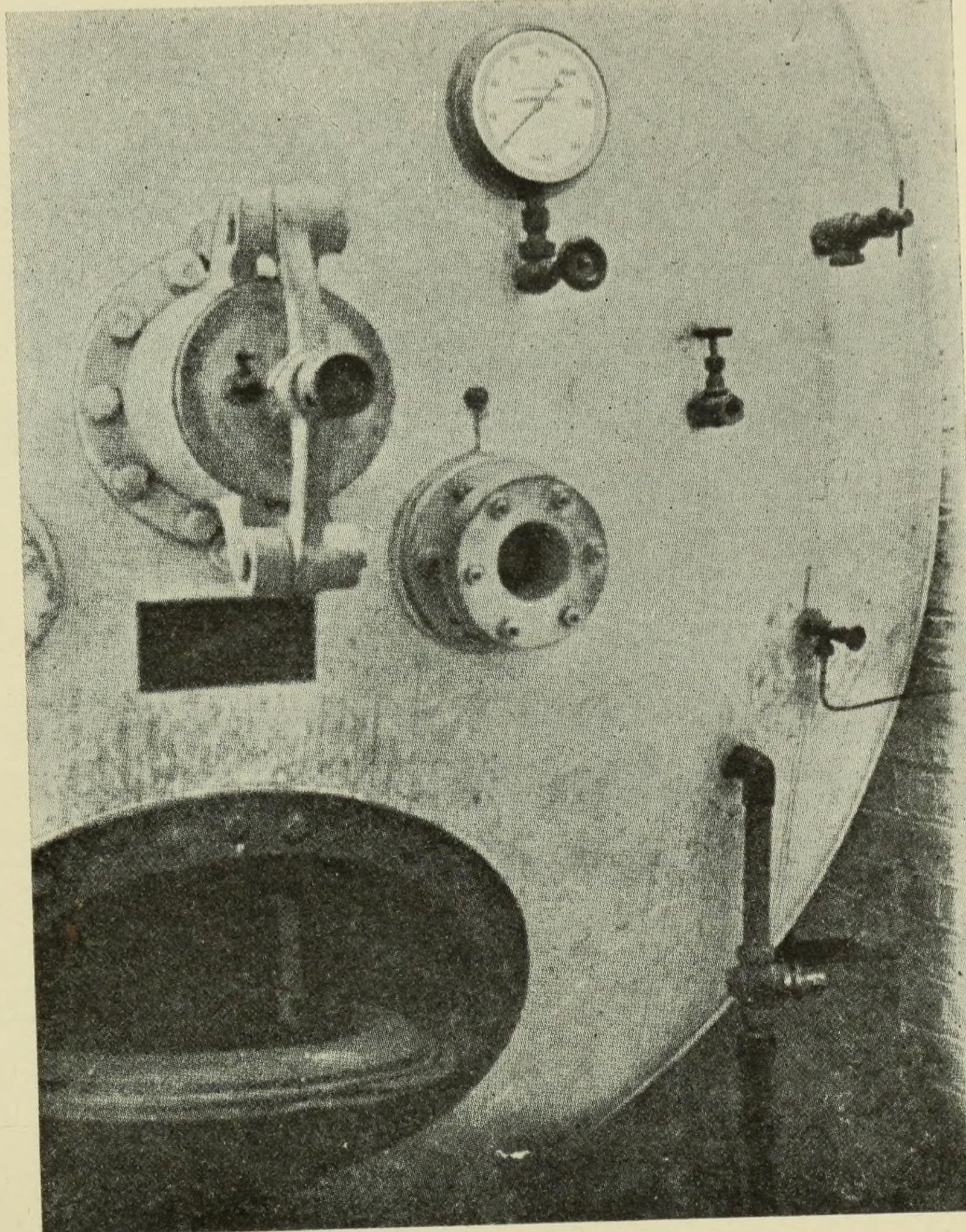 The dished steel end of a large pressure chamber, with an oval manhole at the bottom, a round flanged air-lock with a lever, a small inspection window, a pressure gauge and several valves