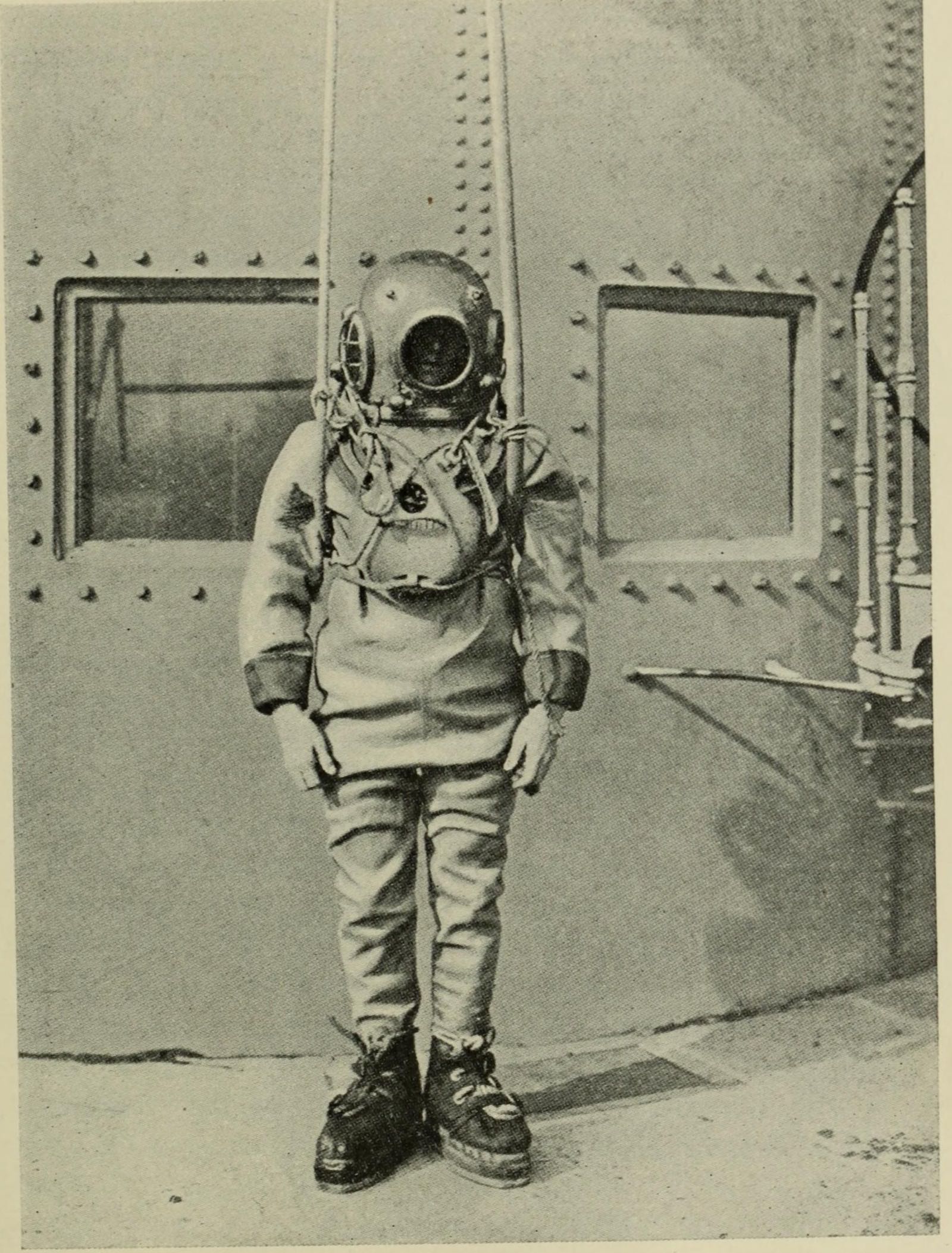 A diver in a canvas suit, copper helmet and heavy boots standing in front of a riveted steel tank, with his air-pipe and life-line running up above him