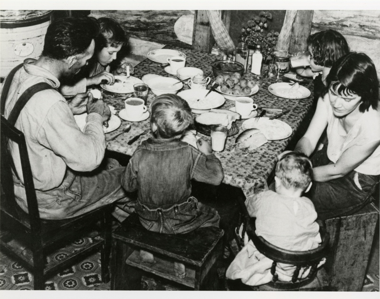 A family of six at a table in a cabin, seen from above, eating from plates with cups of coffee and a glass of milk, a toddler in a high chair at the end