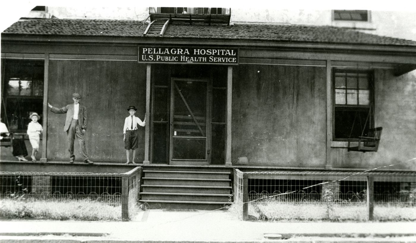 A low frame building with a deep porch and a sign reading Pellagra Hospital, U.S. Public Health Service, with a man and three children standing on the porch