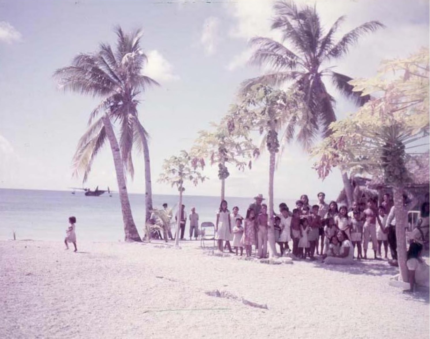People of Utirik standing under coconut palms on a white beach, with a flying boat on the lagoon behind them, in a faded colour photograph