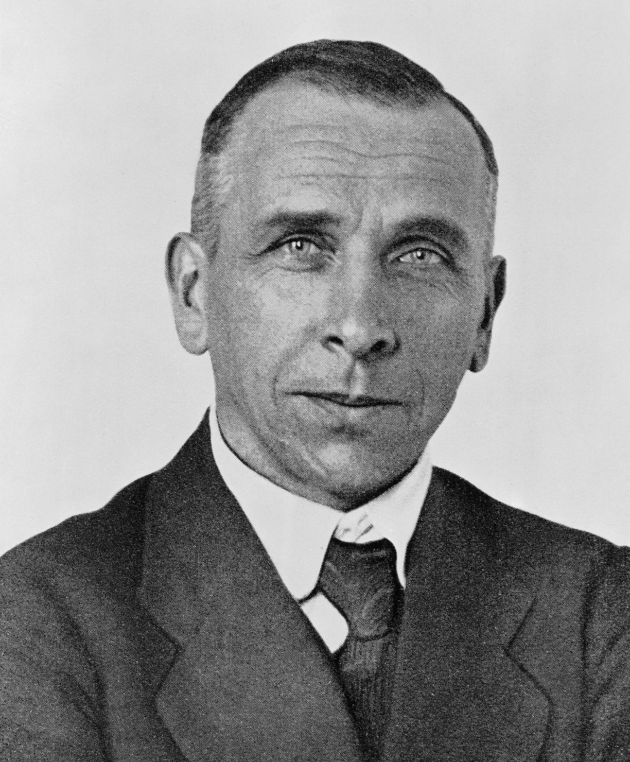 Head-and-shoulders portrait of a man of about forty-five with short hair, a lined forehead and light eyes, in a dark jacket, white collar and tie, looking slightly past the camera