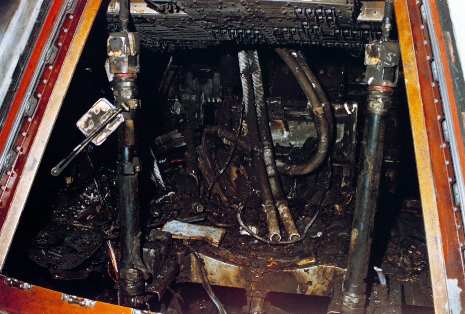 The open hatchway of the command module, framed in charred orange heat-shield material, with the blackened couch struts, burned hoses and melted equipment inside
