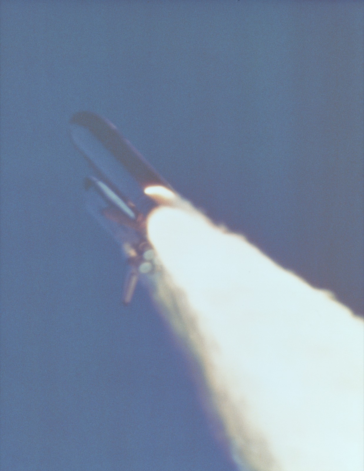 The Shuttle stack seen from below against a blue sky, with a bright plume of flame breaking out of the side of the right-hand booster near its base