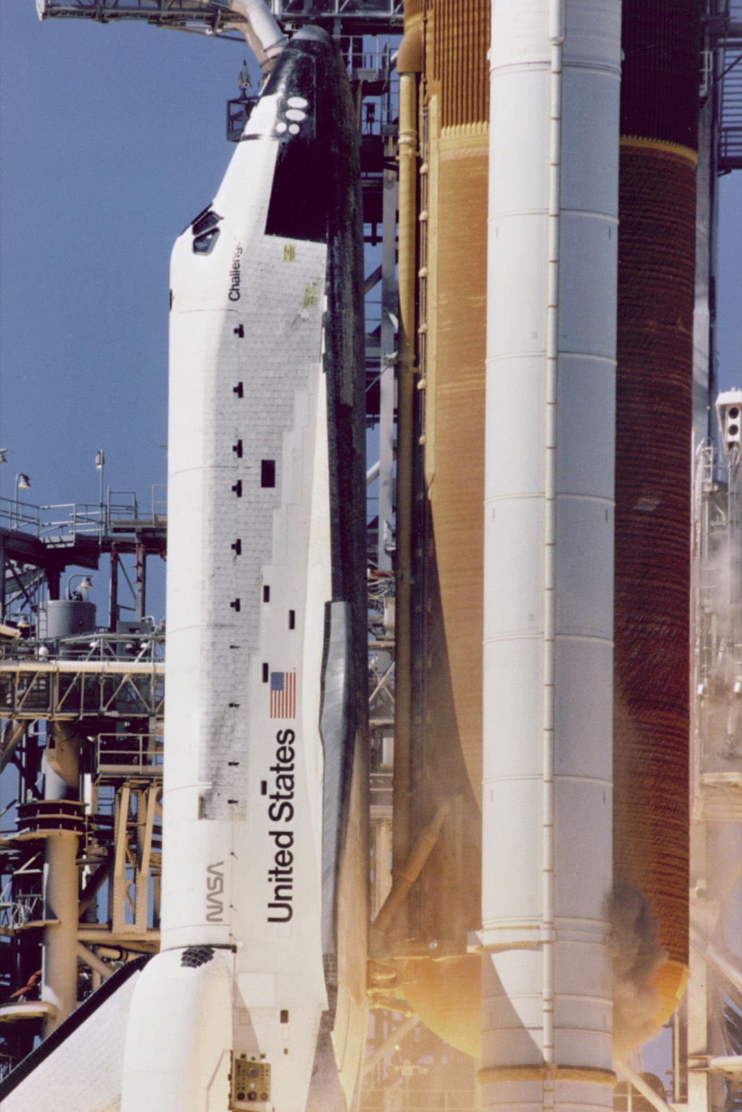 The Space Shuttle Challenger rising past the launch tower, with a small puff of grey-brown smoke on the right-hand booster level with the word United States on the orbiter's side