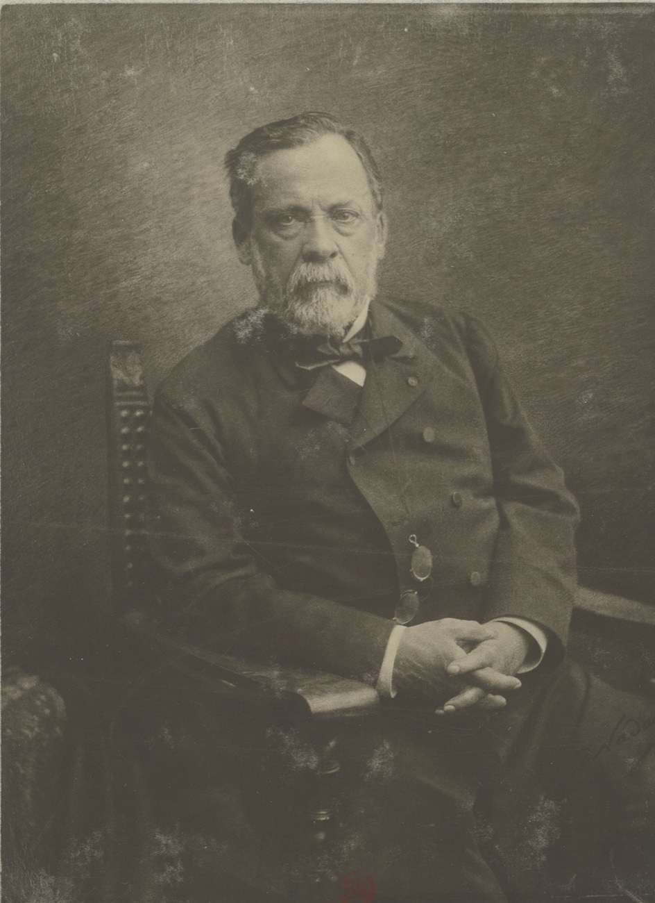A studio photograph of Louis Pasteur seated, grey-bearded, in a dark double-breasted coat with pince-nez on a cord, hands folded on the arm of a chair