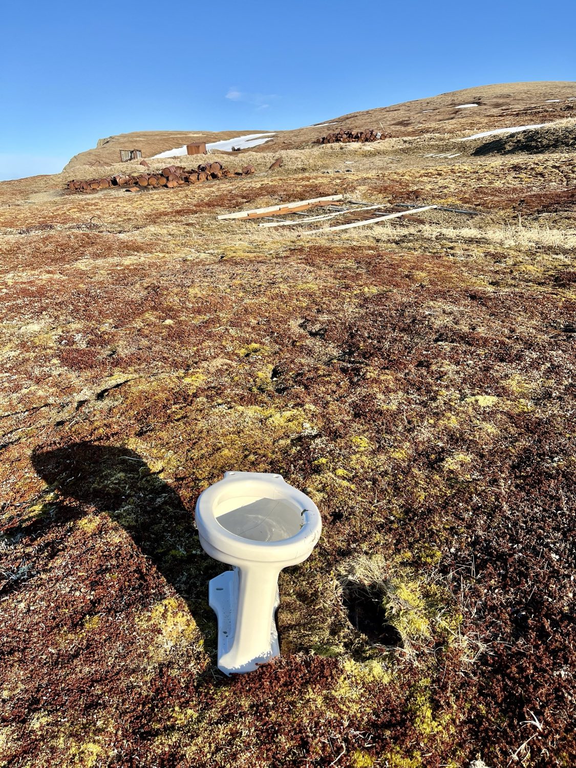 A white porcelain toilet standing alone on brown tundra, with rusted oil drums and the remains of wooden frames on the slope behind