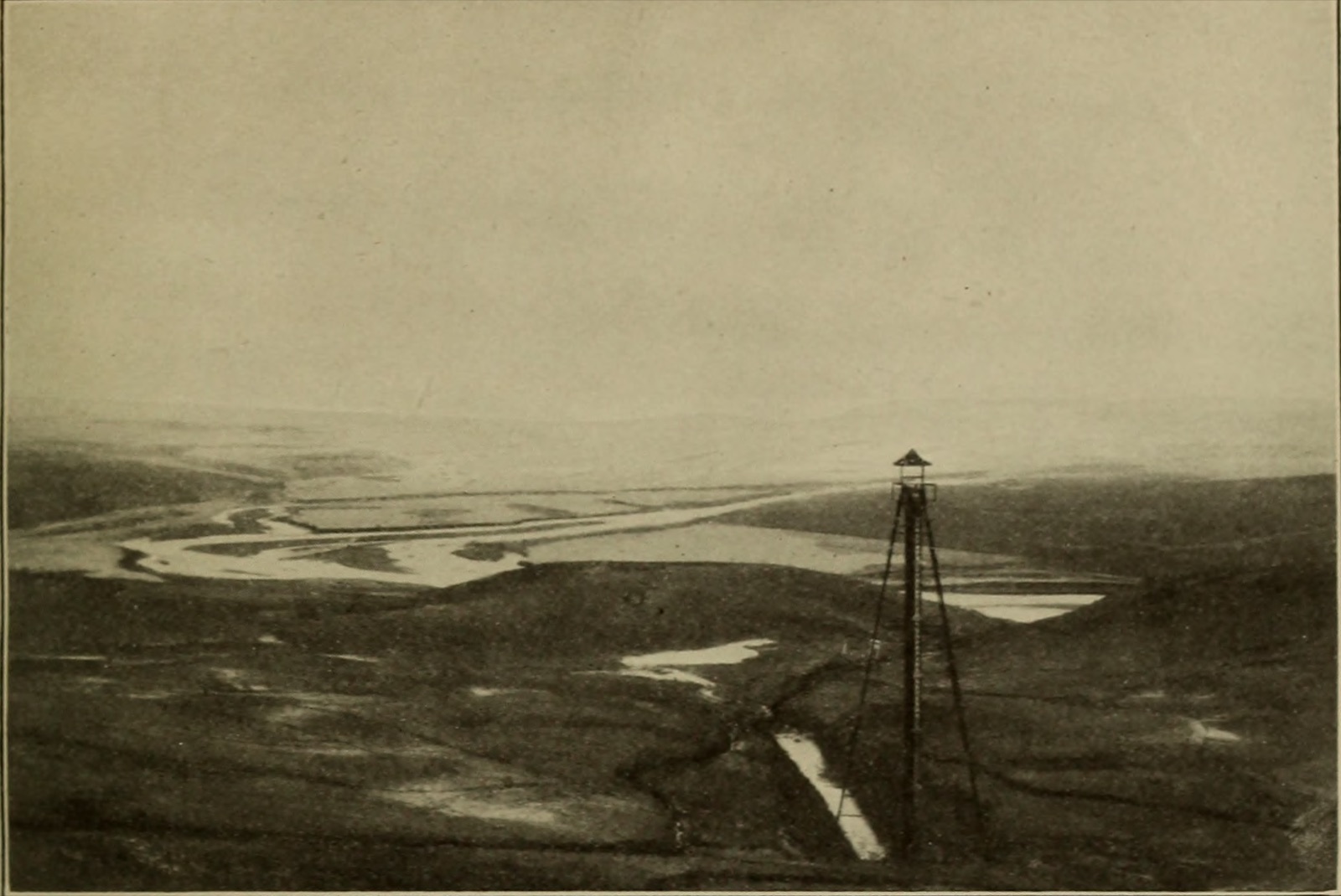 A view over the emptied Lower Otay reservoir, its floor crossed by channels of water, with a tall timber tripod gauge standing in the foreground