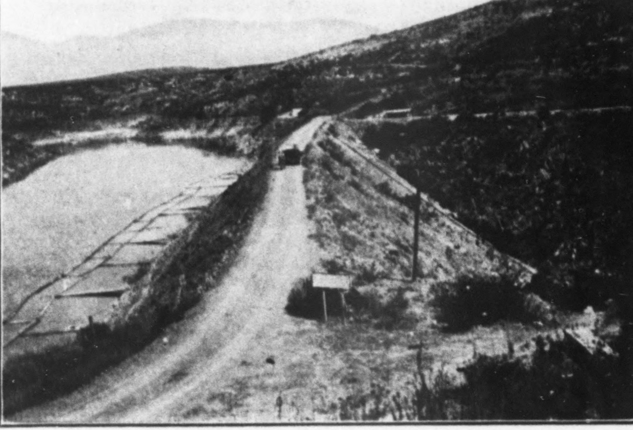 A dirt road running along the crest of a rock-fill dam between hills, with a reservoir on the left, in a halftone photograph