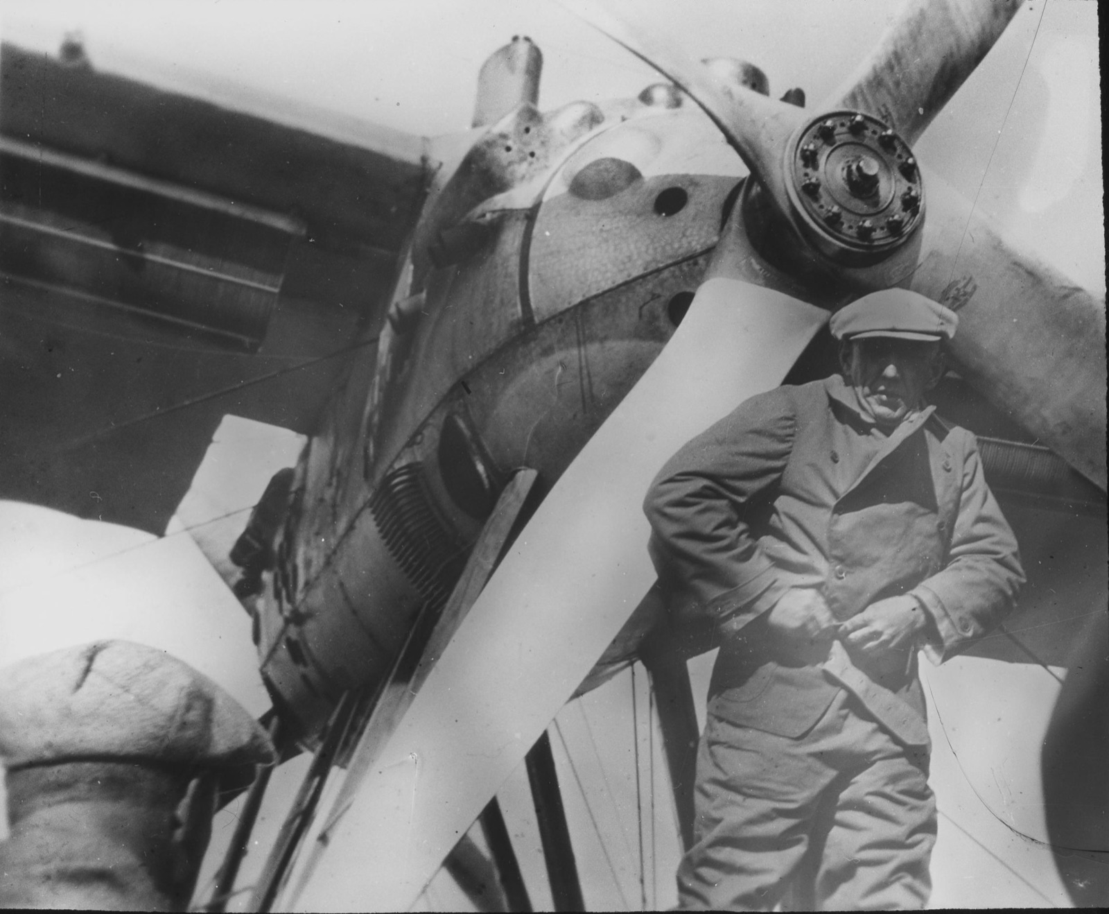 A stocky man in a cloth cap and canvas flying clothes stands under the propeller and engine of a seaplane, squinting at the camera