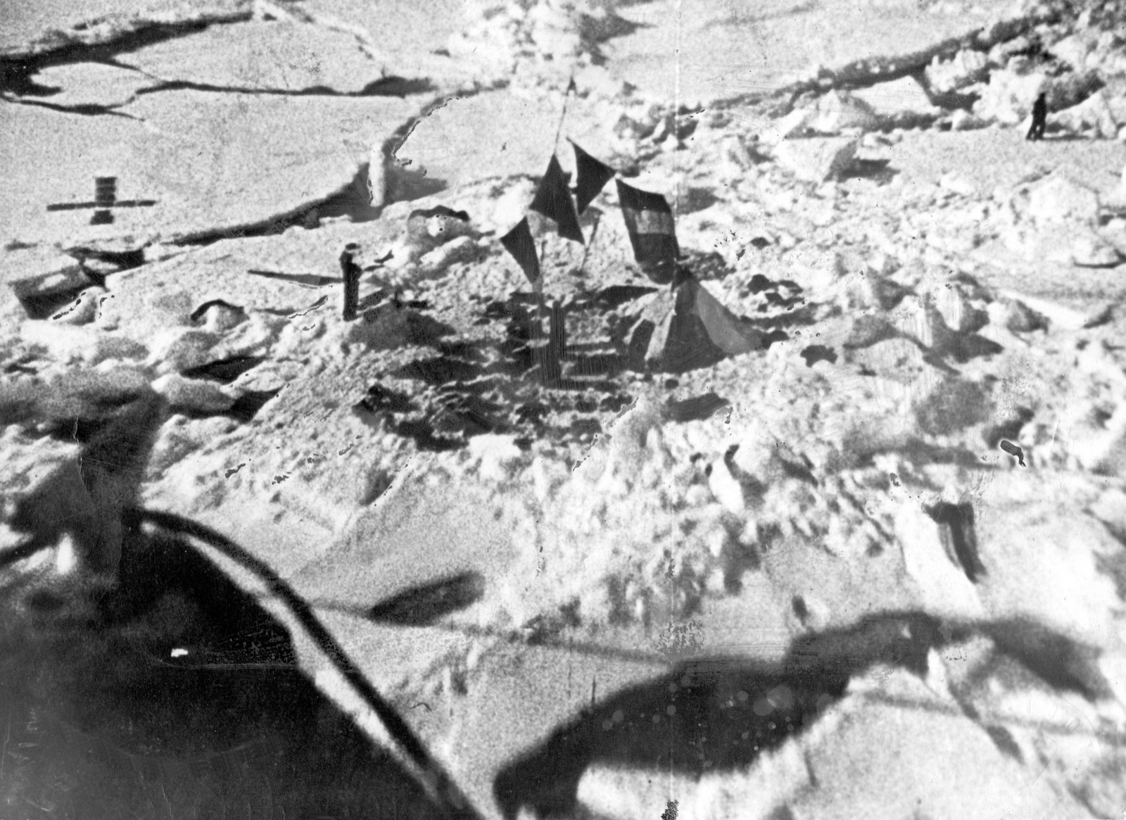 Seen from a low-flying aeroplane with its strut dark in the foreground: a small tent on broken pack ice, flags on a mast beside it, and a lone figure walking at the top right