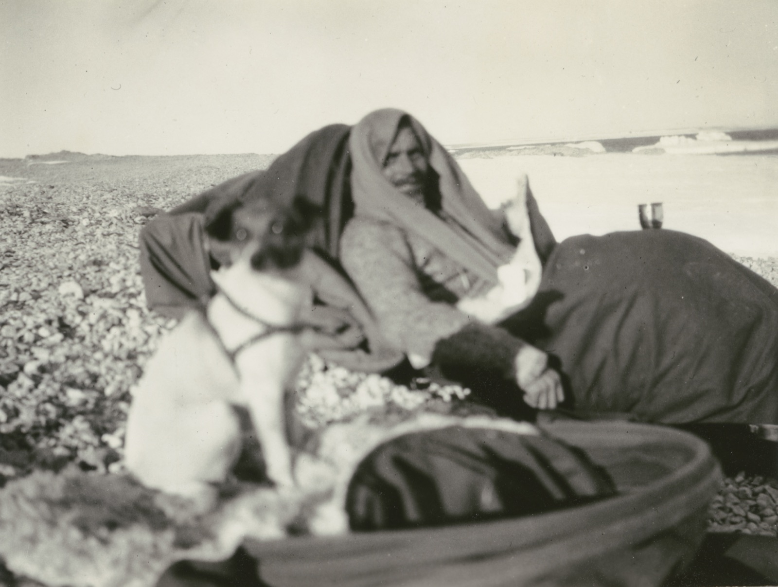 A bearded man wrapped in a blanket sits propped on a stony beach with a small terrier in front of him and ice floes on the water behind