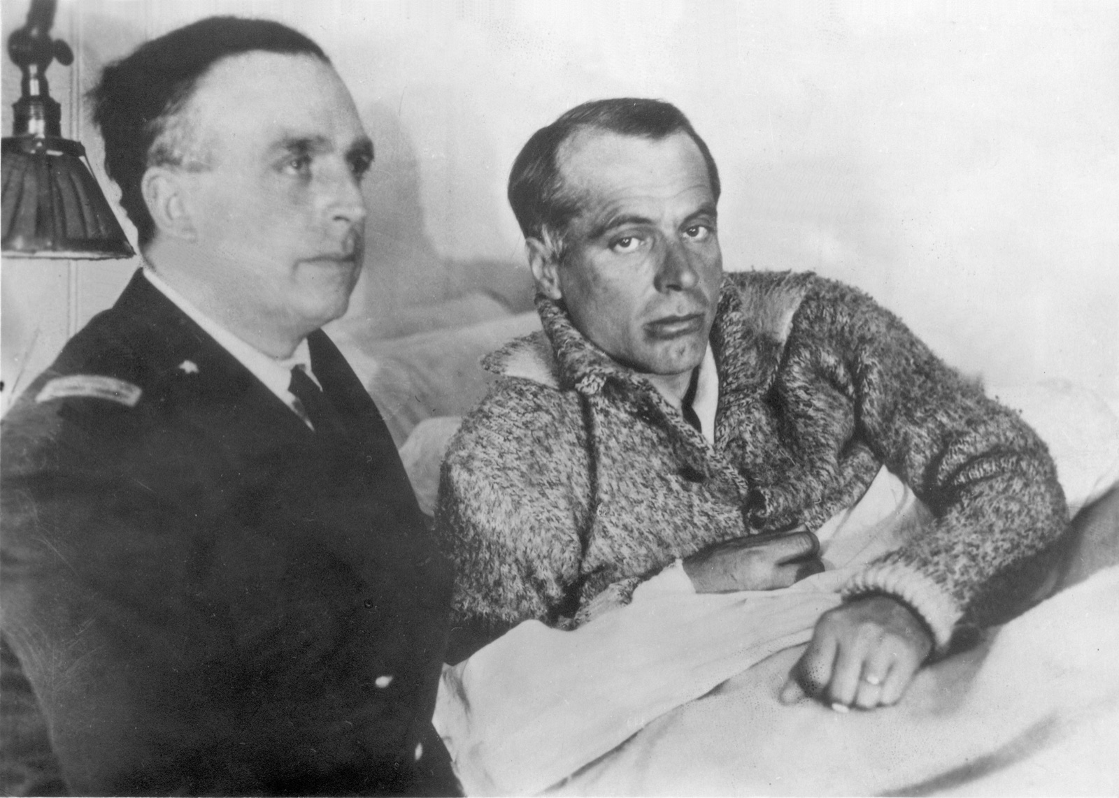 A thin, unshaven man in a thick knitted jersey sits up in a ship's bunk, one hand on the sheet, beside a naval officer in a dark uniform who looks away