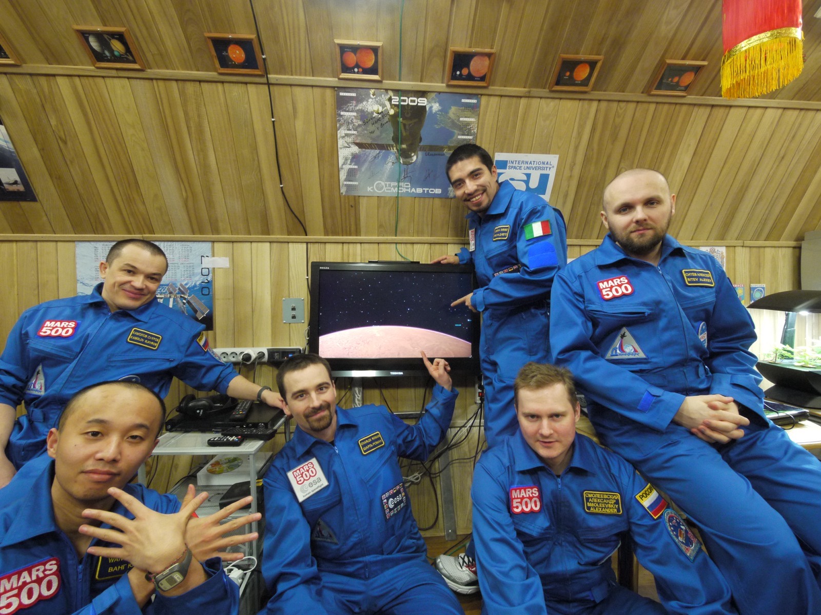 Six men in blue Mars500 overalls with name badges posing in a wood-panelled room around a television showing a simulated view of Mars from orbit