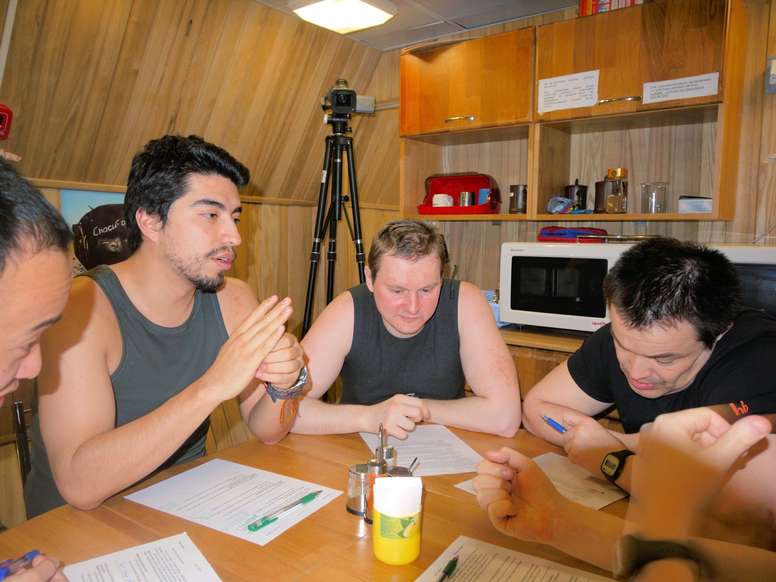 Four of the crew at the kitchen table in a wood-panelled module, filling in printed questionnaires with pens, with a video camera on a tripod behind them