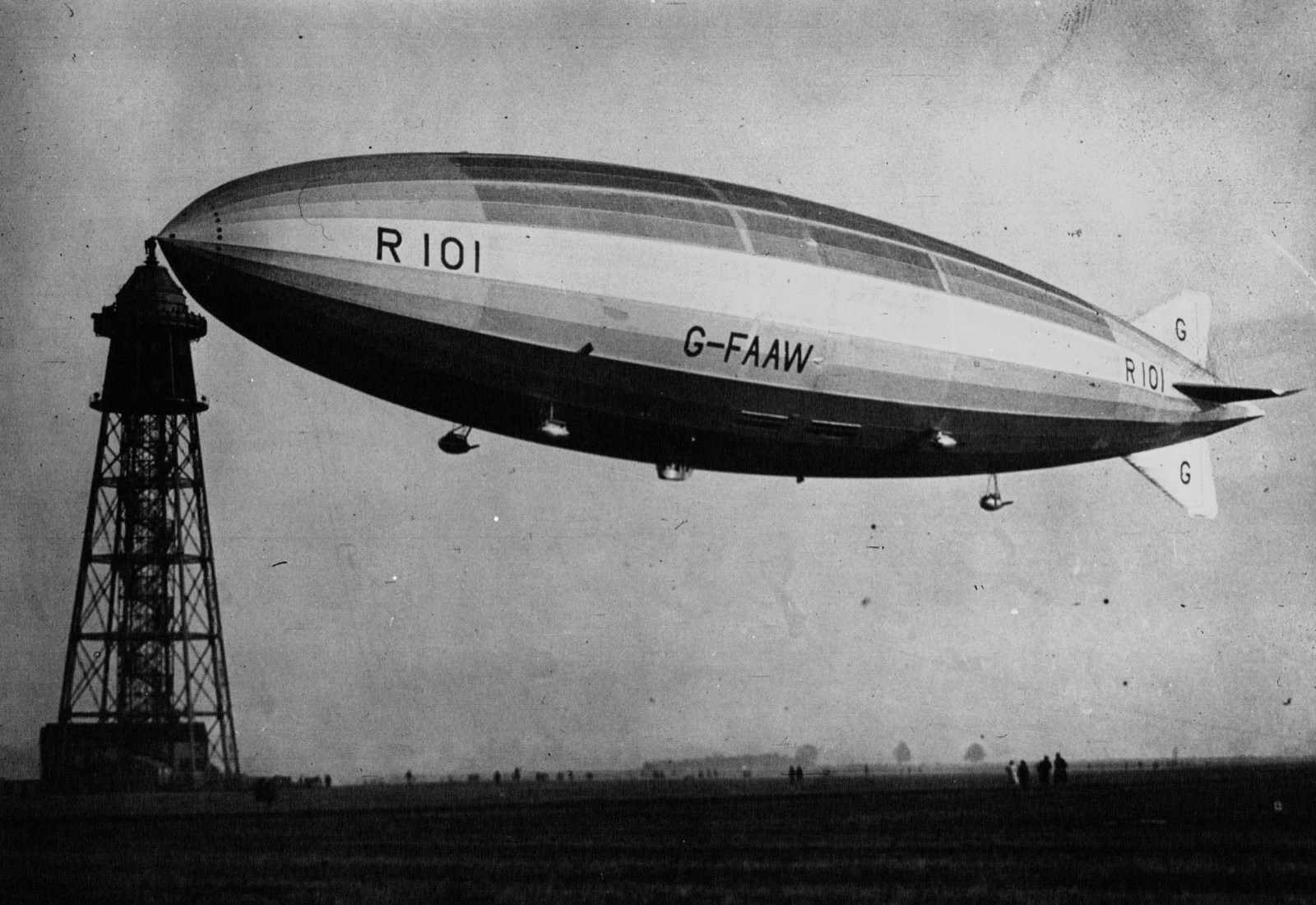 A long silver airship with R101 and G-FAAW painted on her side hangs from the top of a tall lattice mooring tower on a flat field, with small figures on the grass below
