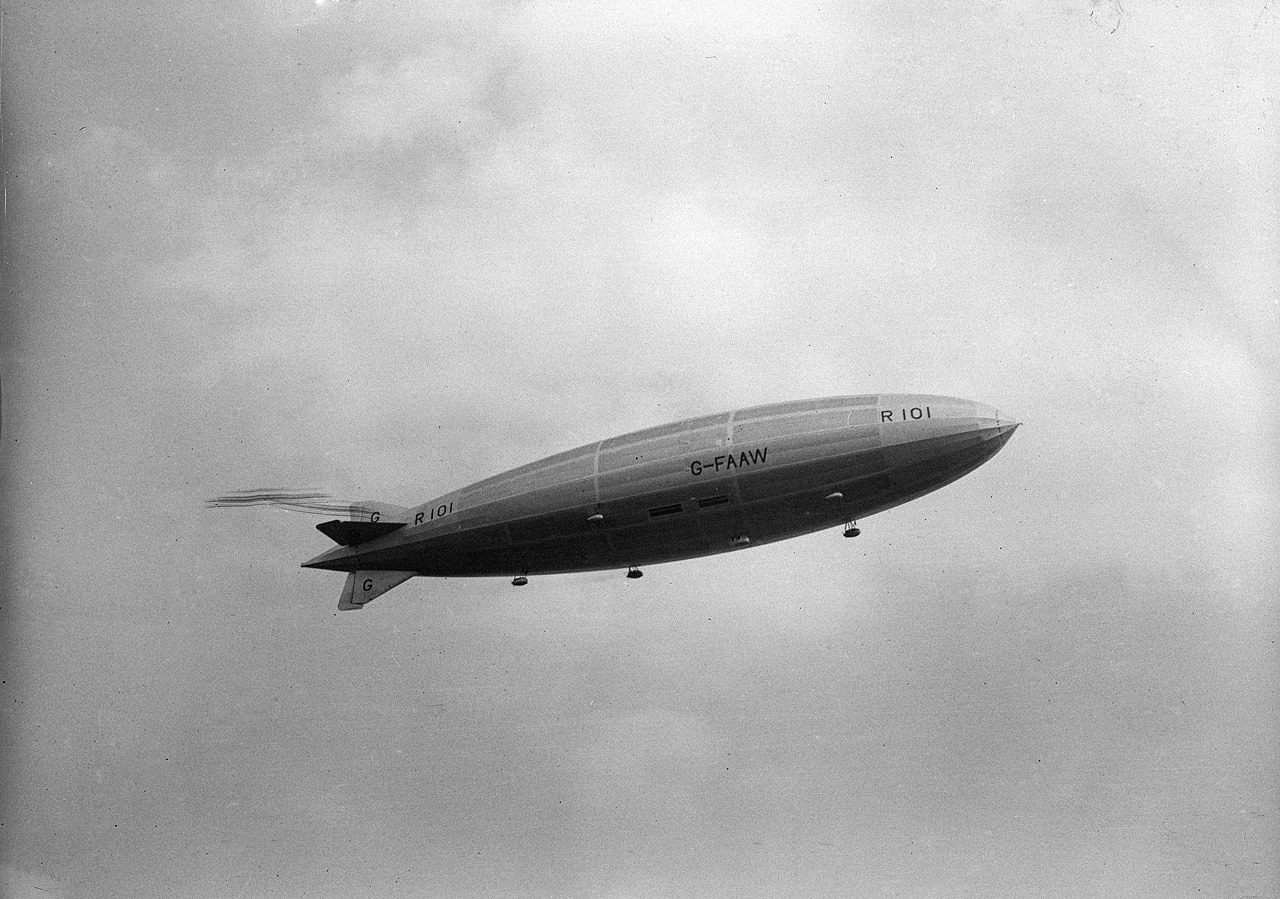 The airship R101 low in a hazy sky over the roofs and chimneys of a suburban street, with people in the road looking up and parked cars at the kerb
