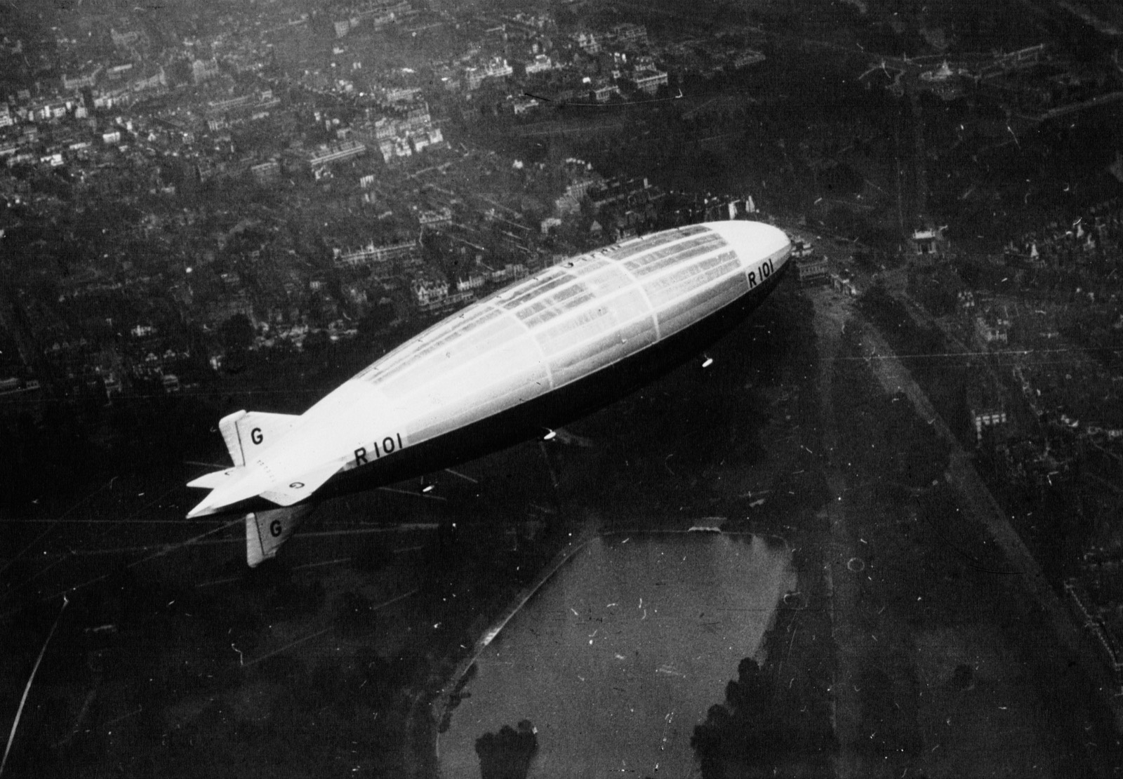 An airship seen from above against the dark roofs and parks of a city, a lake with islands beneath her tail