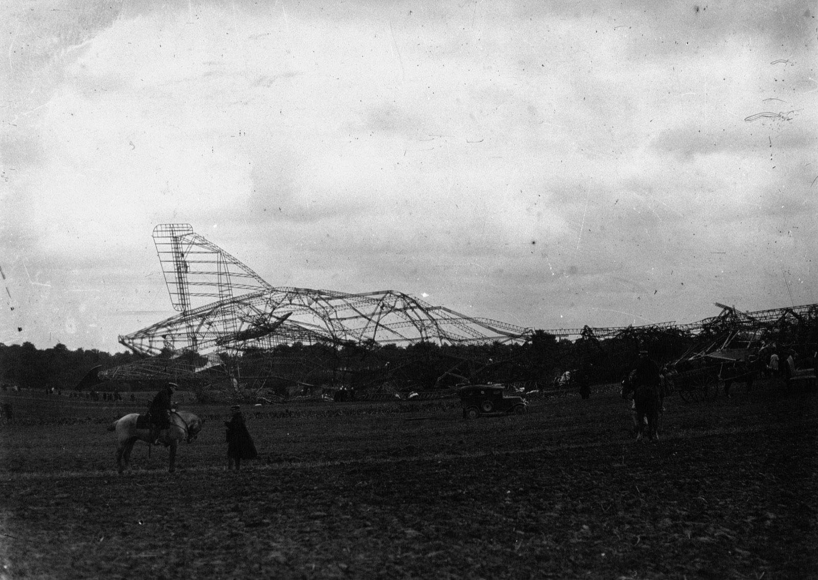 The bare metal skeleton of an airship lies across a field with its tail fin standing up at the left, a car and a cart beside it and two mounted men in the foreground