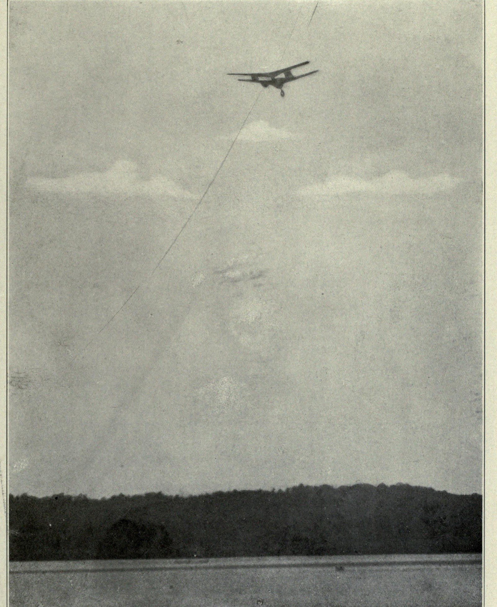 A small tandem-winged machine high in a grey sky above a wooded shoreline, photographed from the water