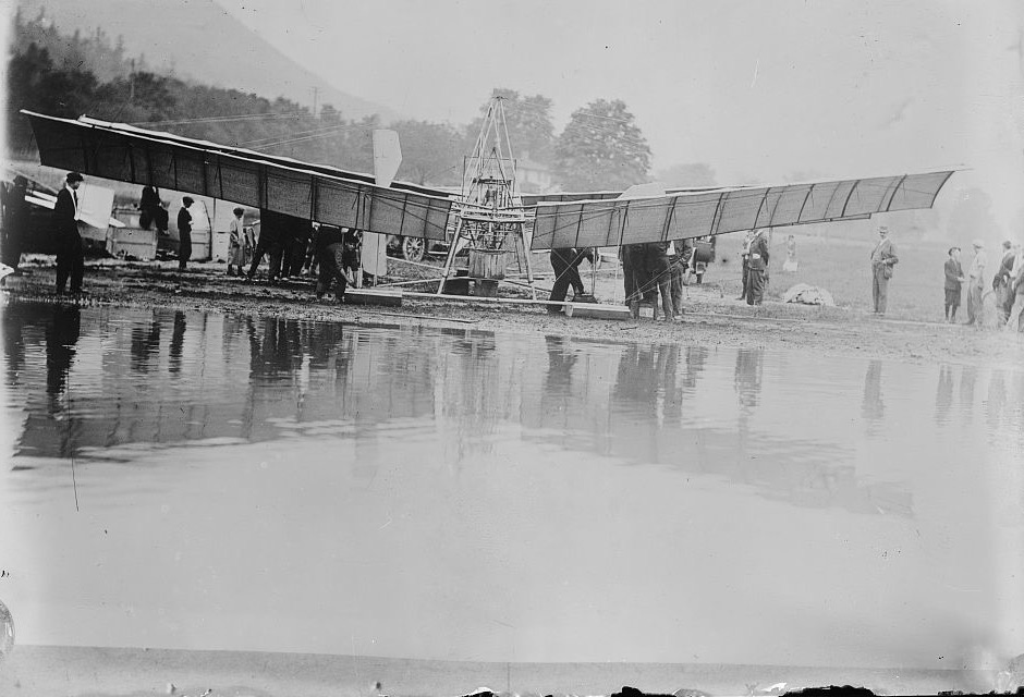 A tandem-winged aeroplane on floats standing at the edge of a lake with a crowd of men and women around it and wooded hills behind