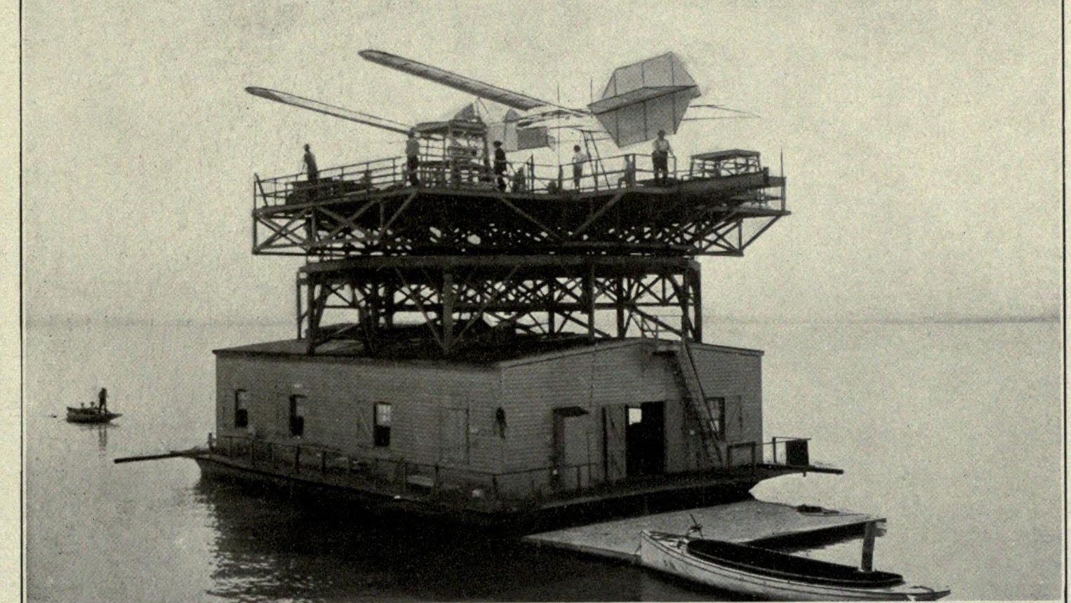 A large two-storey houseboat on calm water carrying a timber superstructure, with a model aerodrome on a track on its top platform and men standing on the platform