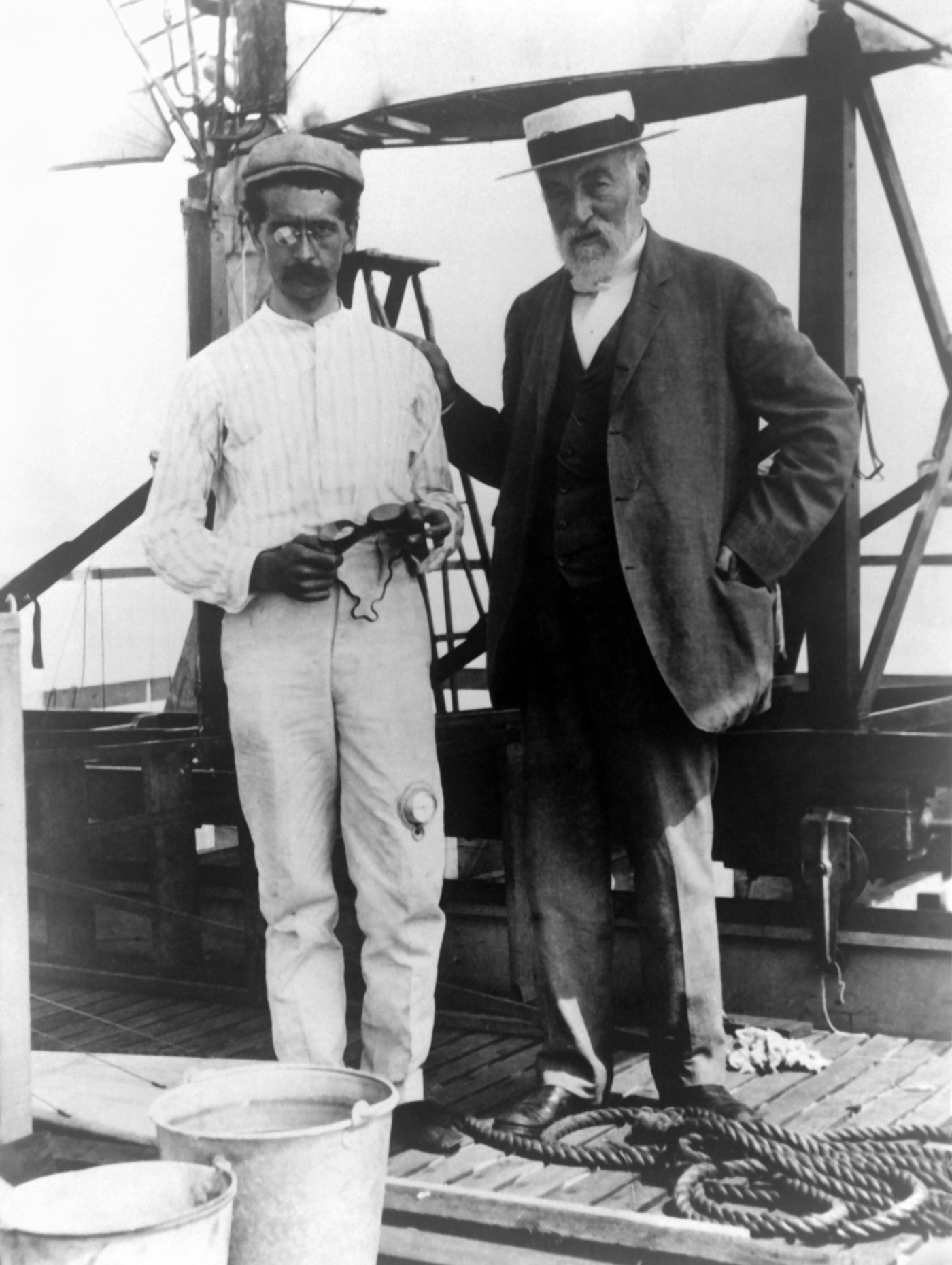 Two men standing on the deck of a houseboat under a timber superstructure: a young man in a striped shirt and cap holding a pair of goggles, and an older bearded man in a dark suit and straw boater with a hand on his shoulder