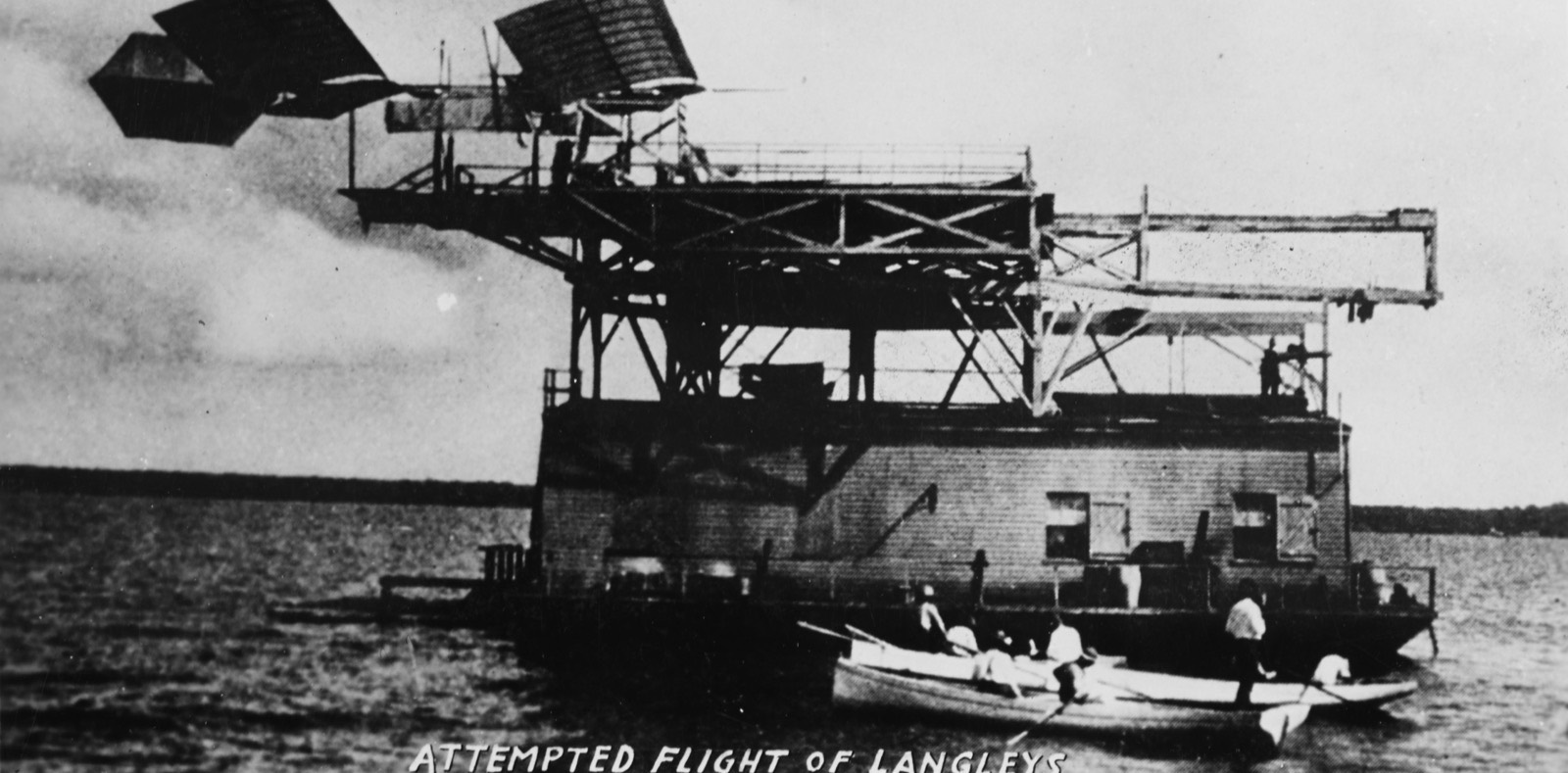 The Aerodrome in the air just beyond the end of the launching track on the houseboat roof, nose down, its front wings twisted, with a rowing boat on the water below