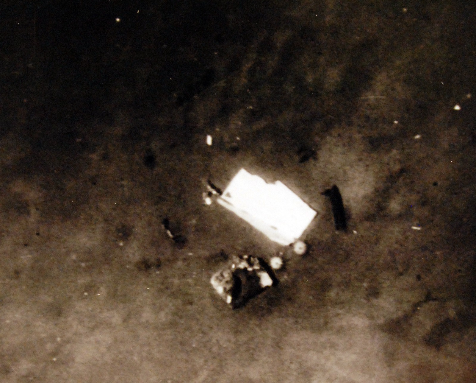 A grainy photograph of the sea floor from above, with a pale rectangular sheet of metal, a dark twisted object and small round shapes on the mud