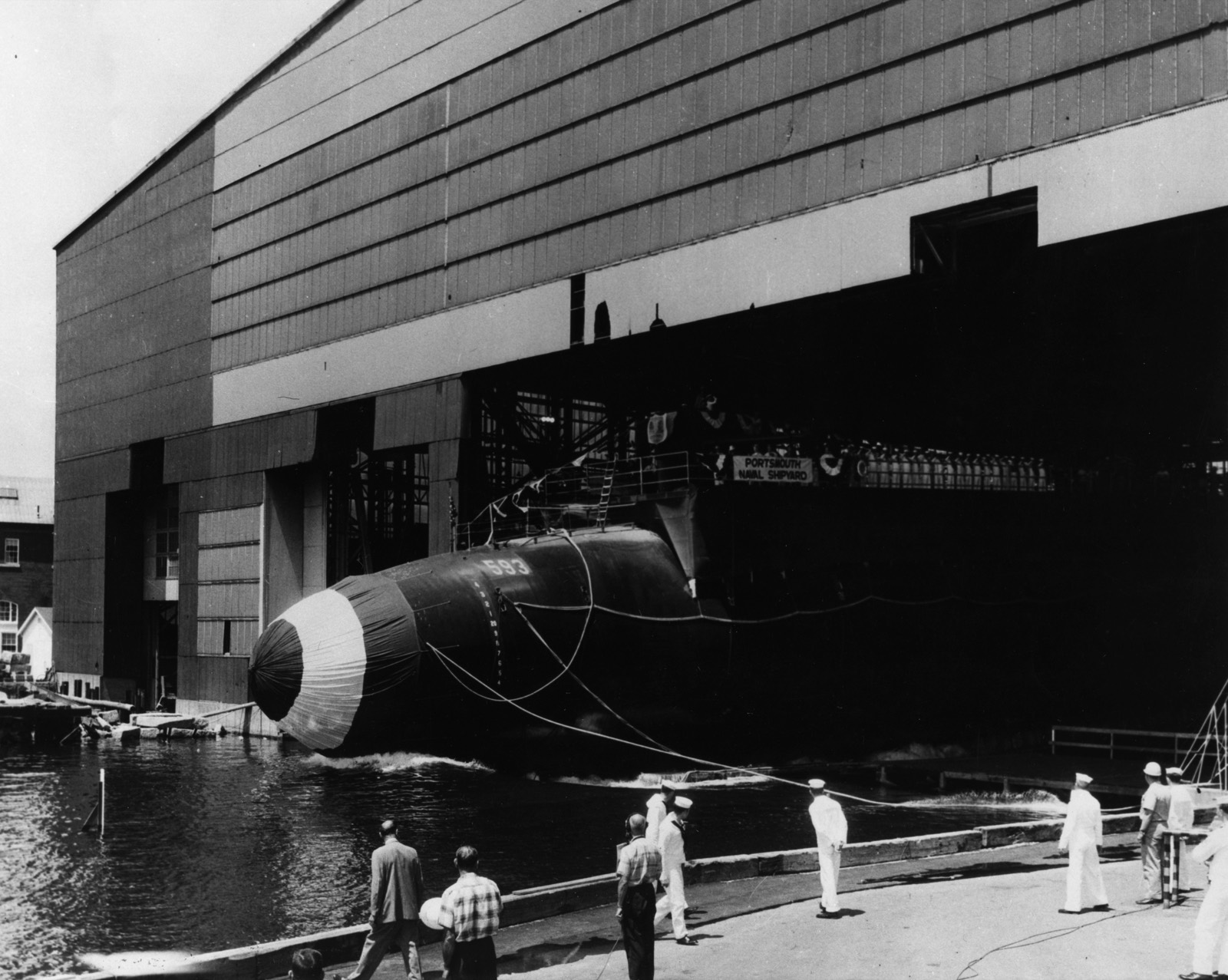 A black submarine hull with a striped bow slides bow-first out of a tall shipbuilding shed into the river while a few men in white uniforms watch from the quay