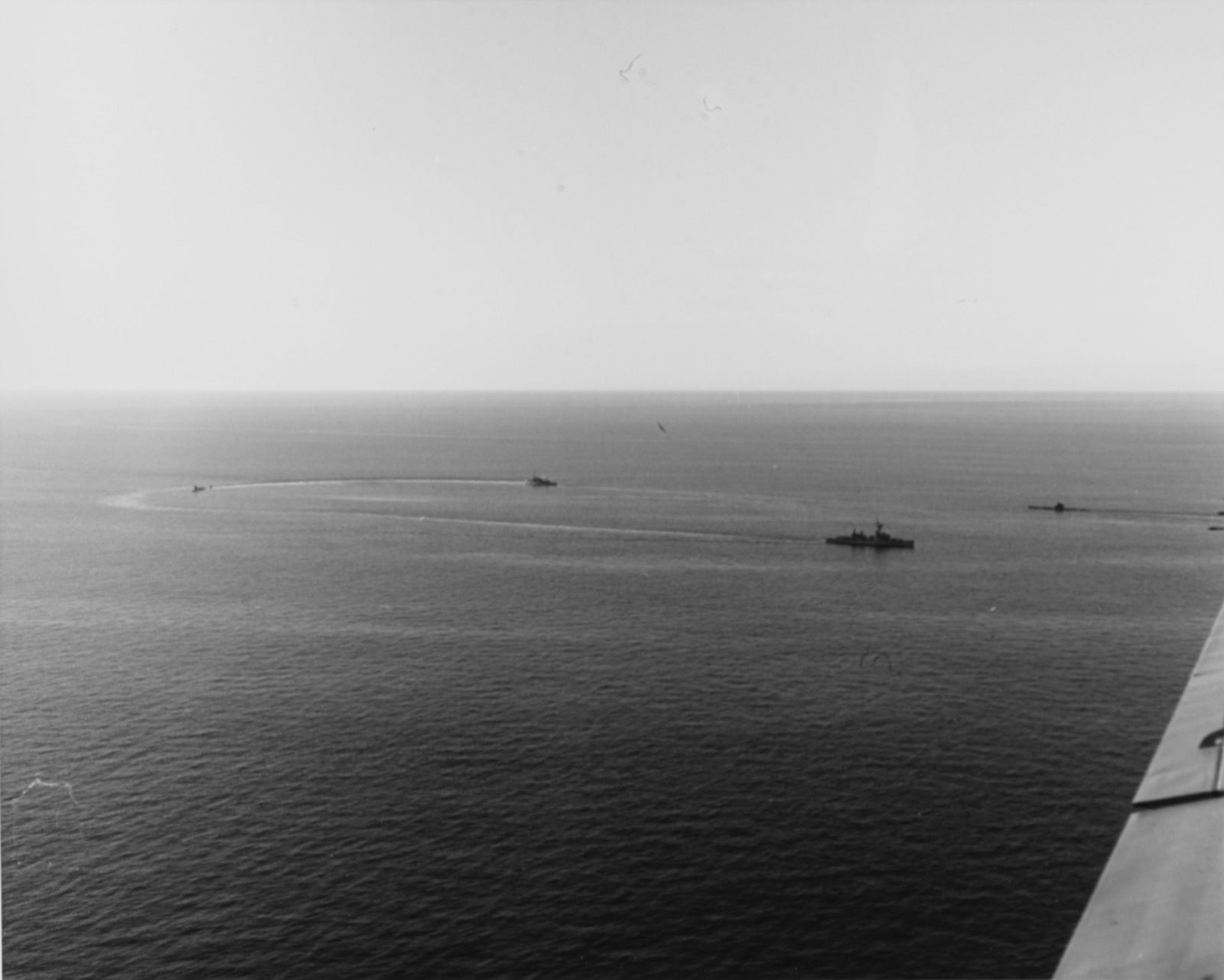 Seen from the air, three ships and a submarine circle on a flat grey sea, the wake of one curving round the others