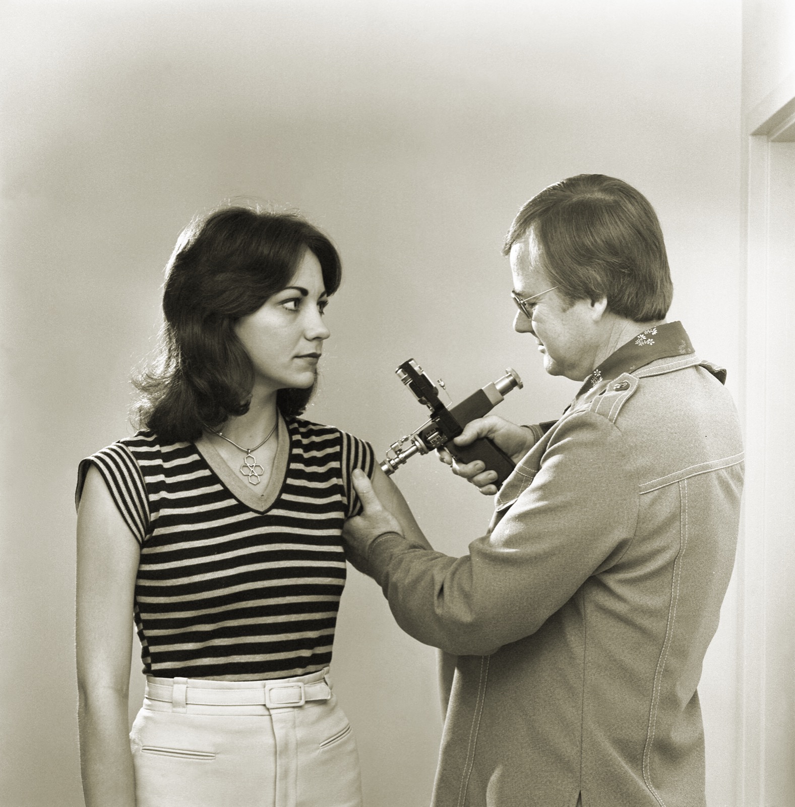 A public health clinician in a 1970s jacket holds a pistol-shaped jet injector against the upper arm of a young woman in a striped shirt, who looks at him