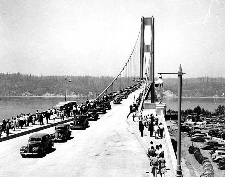 Opening day on the bridge: a column of 1940 cars and a crowd of people on foot fill the two-lane deck, looking west along the cables to the towers and the far shore, with lamp standards on the kerbs and the water on either side