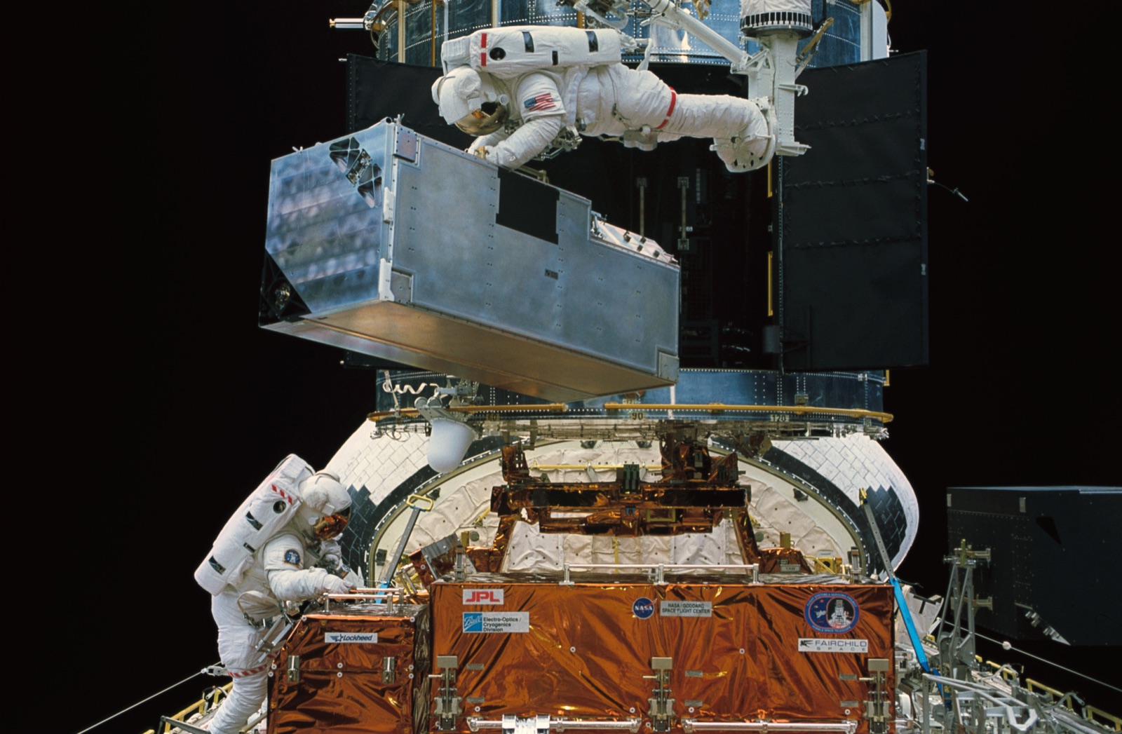 An astronaut on the end of the shuttle's arm holds a large grey instrument box above the open bay of the telescope while a second astronaut works at the foot of the bay