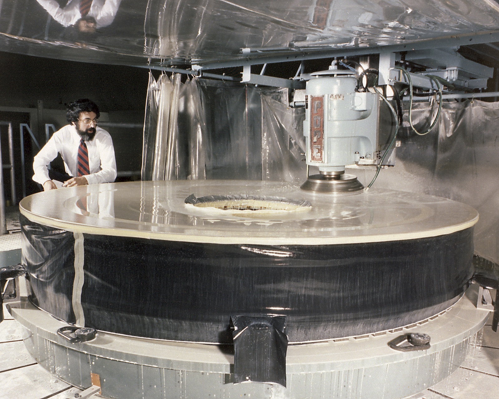 A man in shirt and tie watches a grinding head at work on the broad grey disc of the primary mirror, which fills a round steel mount under plastic curtains