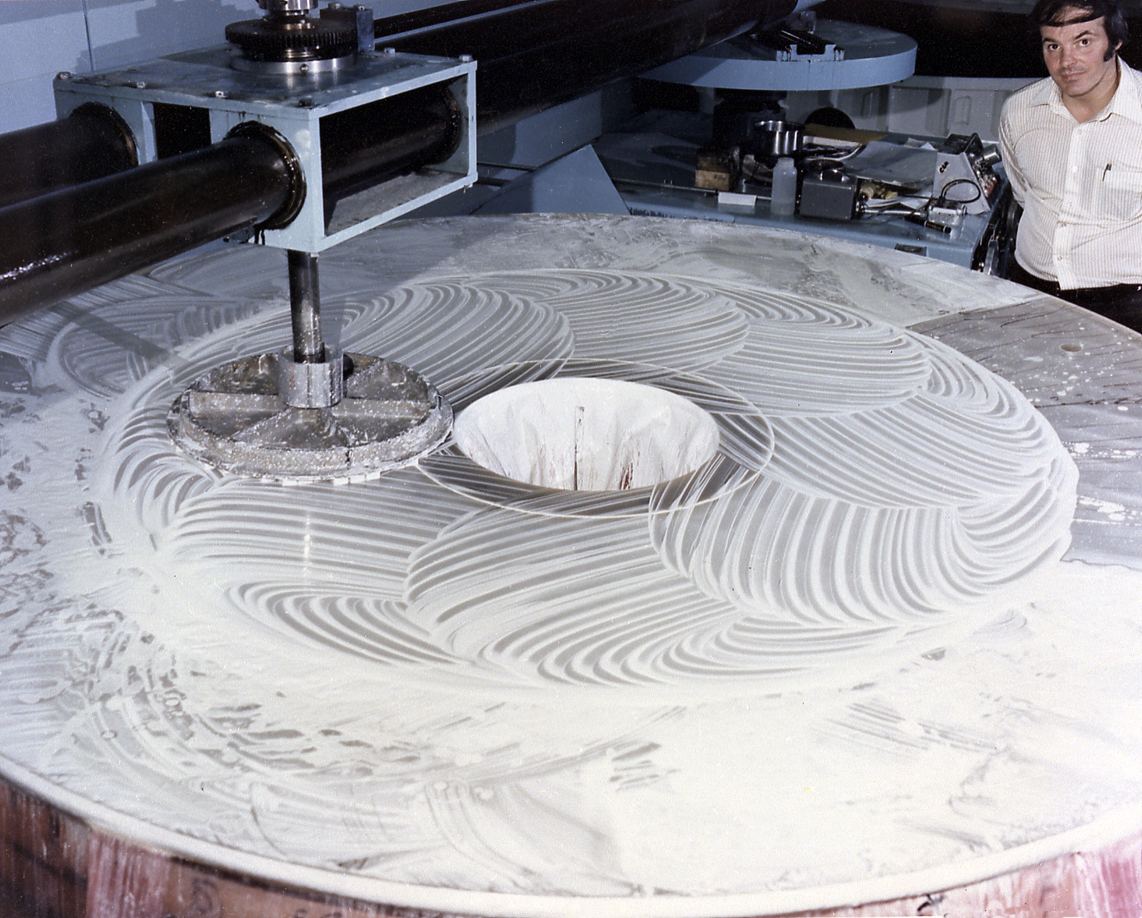 The primary mirror face up, its surface covered in white polishing compound swirled into overlapping arcs by a small tool on an arm, with a technician watching from behind