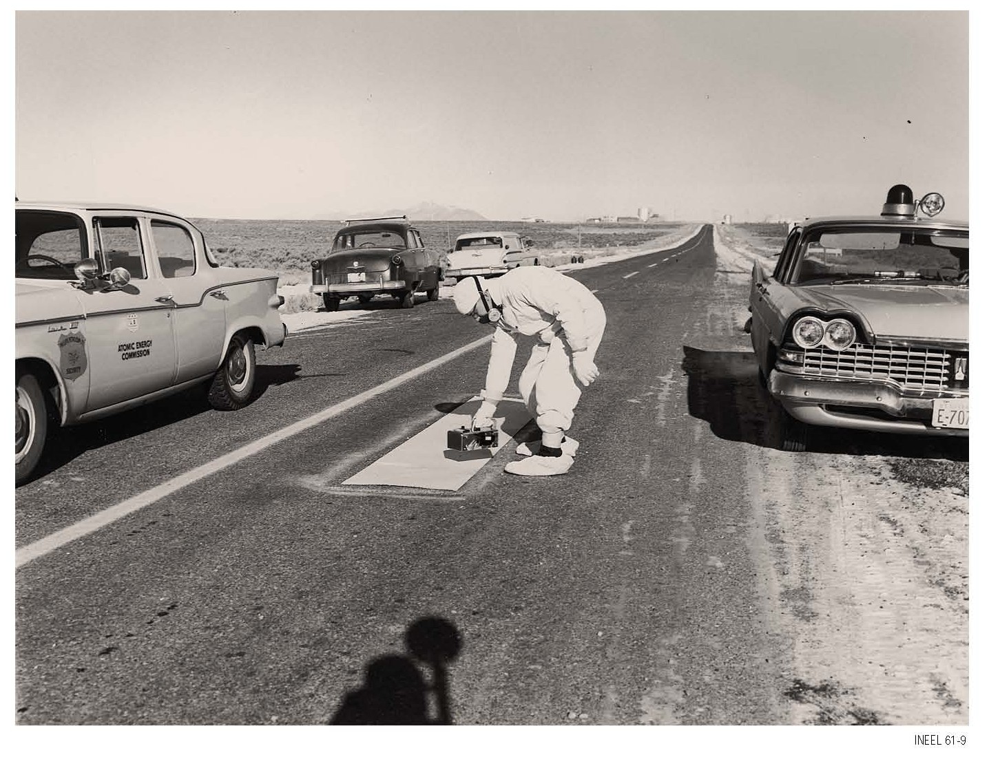A man in white protective clothing bends over an instrument on a sheet laid on a two-lane desert highway, between an Atomic Energy Commission station wagon and a police car, with mountains far off