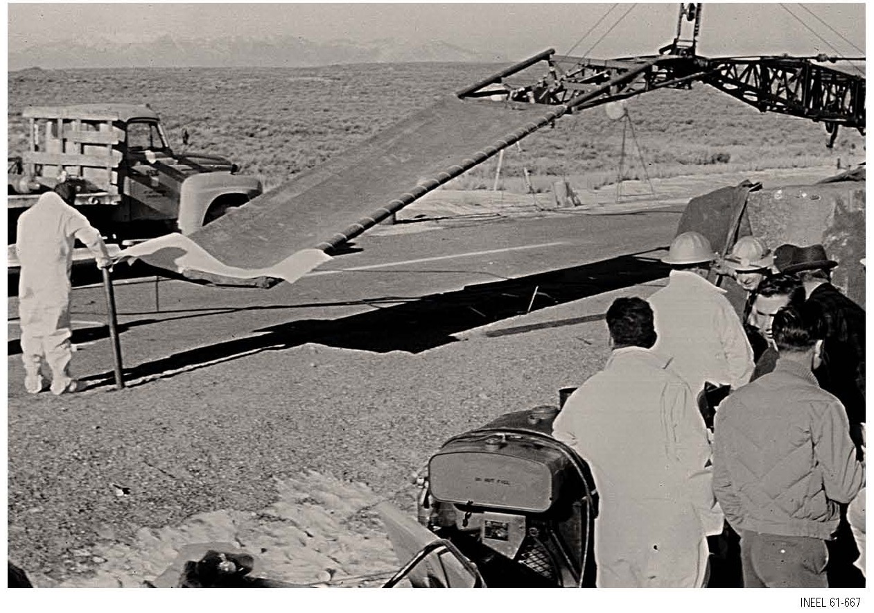 Men in hard hats and white suits stand on gravel beside a long netted stretcher rig hanging from a crane boom, with a flatbed truck and sagebrush desert behind