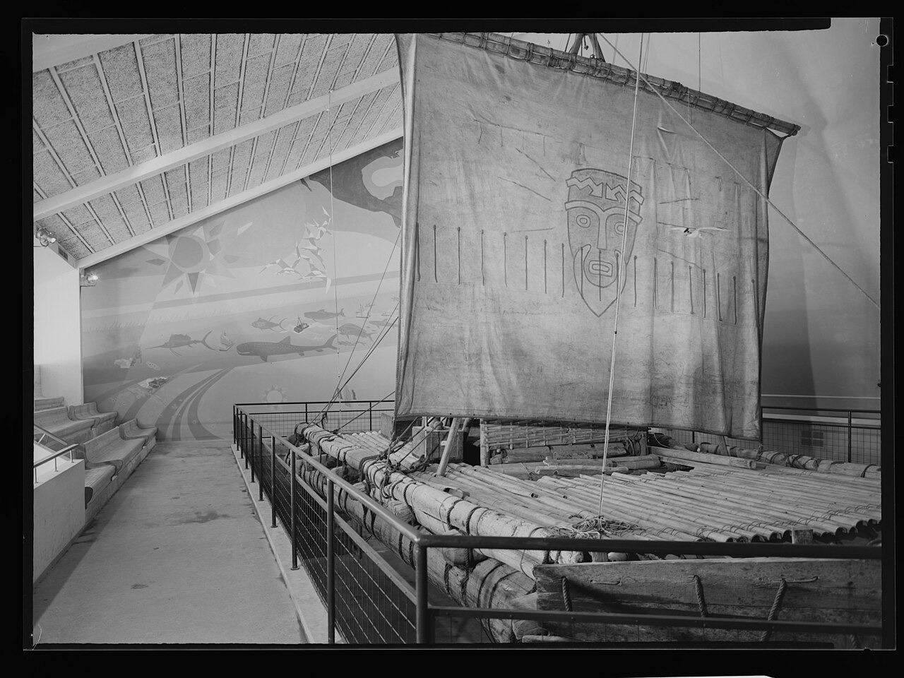 The raft in a museum hall, its square sail raised and painted with a bearded sun-god's face, bamboo deck and lashed logs behind a railing, a mural of fish and a sun on the wall beyond