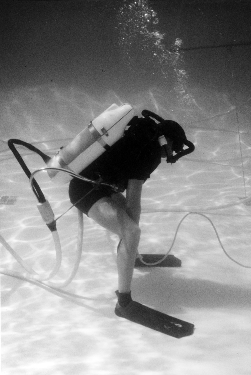 An underwater photograph of a diver in a wetsuit, fins and mask bent forward in shallow sunlit water, a white cylinder on his back and hoses trailing to a surface umbilical
