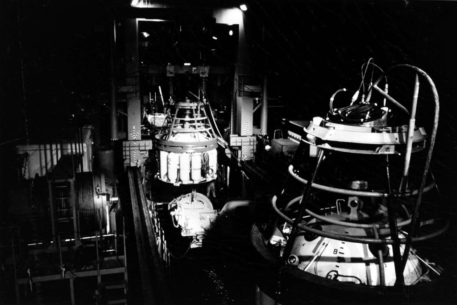 Inside a dim ship's well, a spherical capsule hangs from a gantry over the water while another capsule, mated to a chamber, stands in the foreground with cables coiled on top