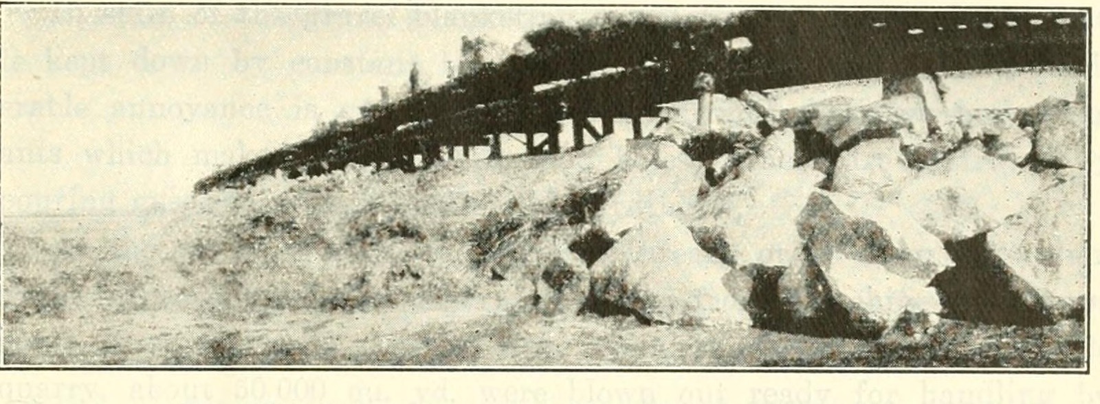 A halftone photograph of a railway trestle on piles with a train of cars standing on it, and large angular blocks of rock piled below it to the right