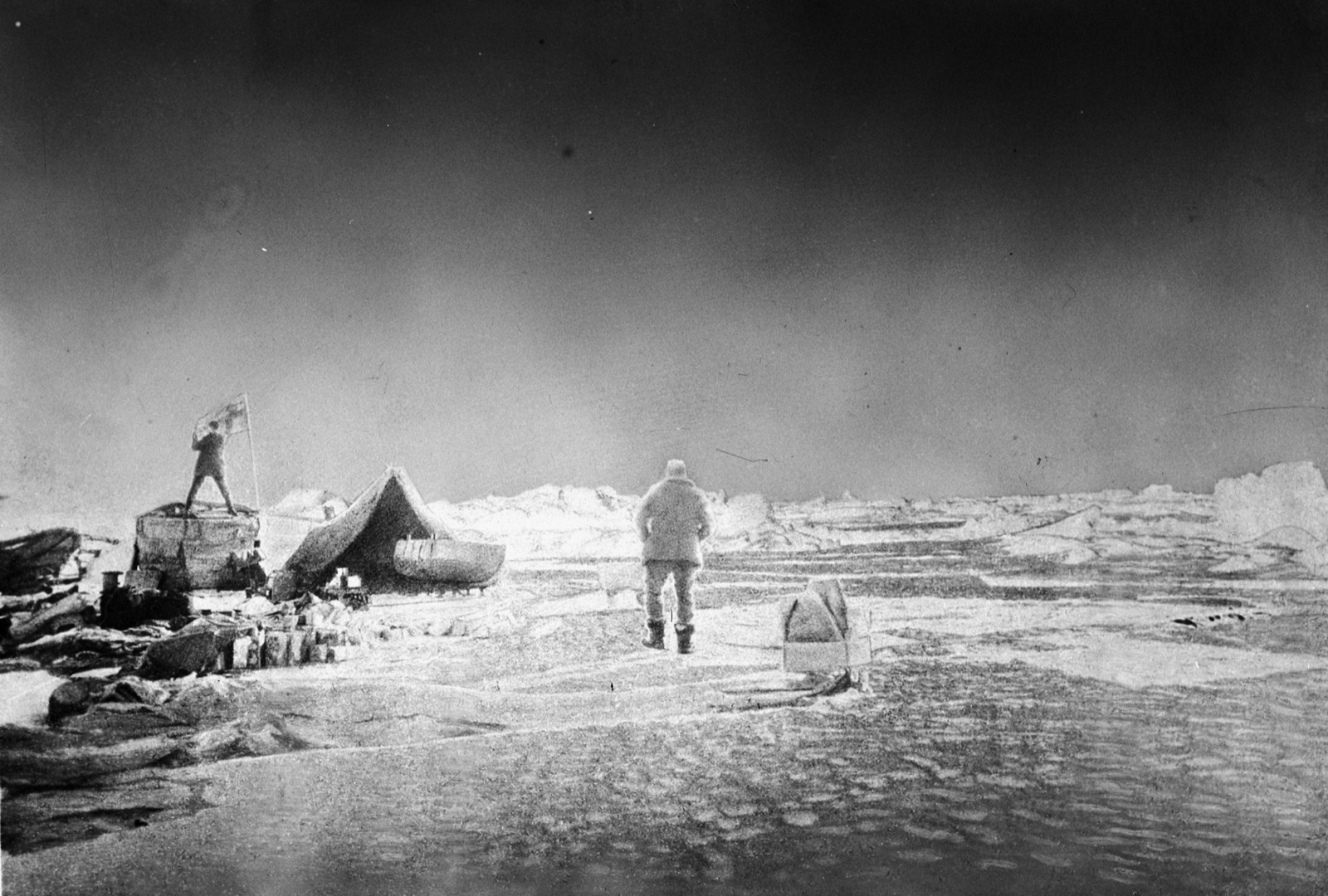 Bright overexposed photograph of the camp on the ice: a man stands on the roof of the balloon car holding a flag, a tent and the canvas boat beside it, another man walking away across the floe with his back to the camera