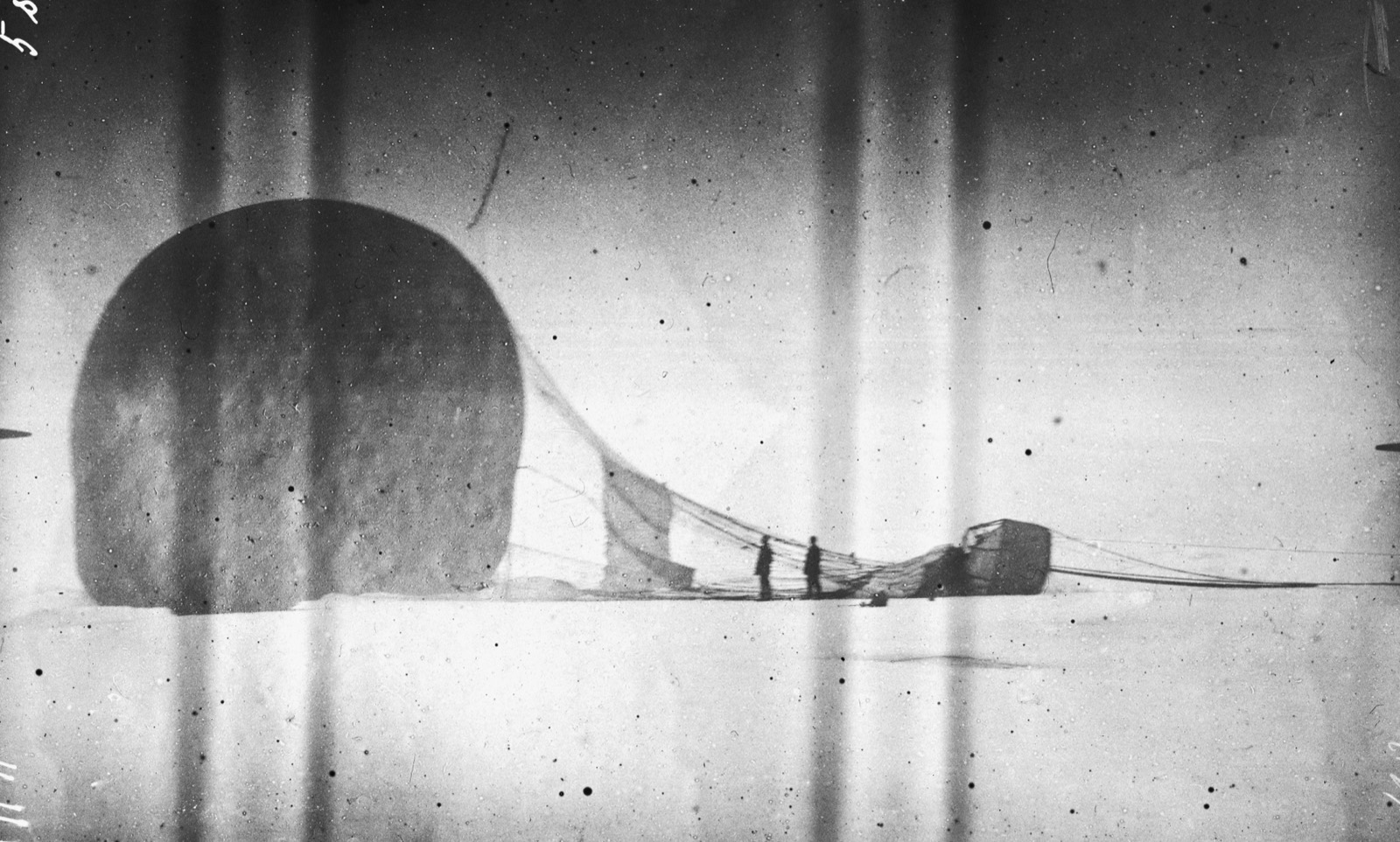 Streaked photograph of a deflated balloon slumped on the ice like a dark dome, its netting trailing to a toppled car, with two small figures standing between them